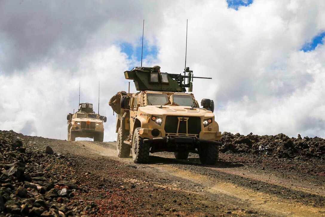 Two military vehicles drive down a dirt road.