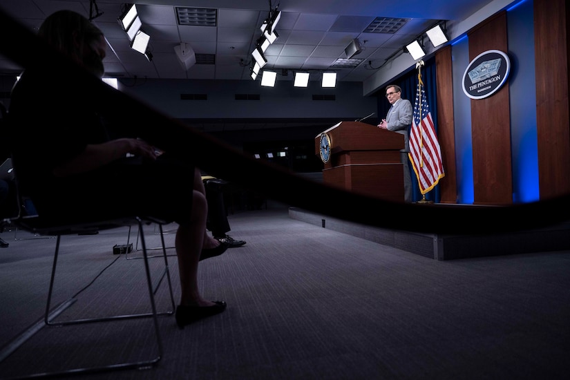 A civilian stands and speaks in front of an American flag.