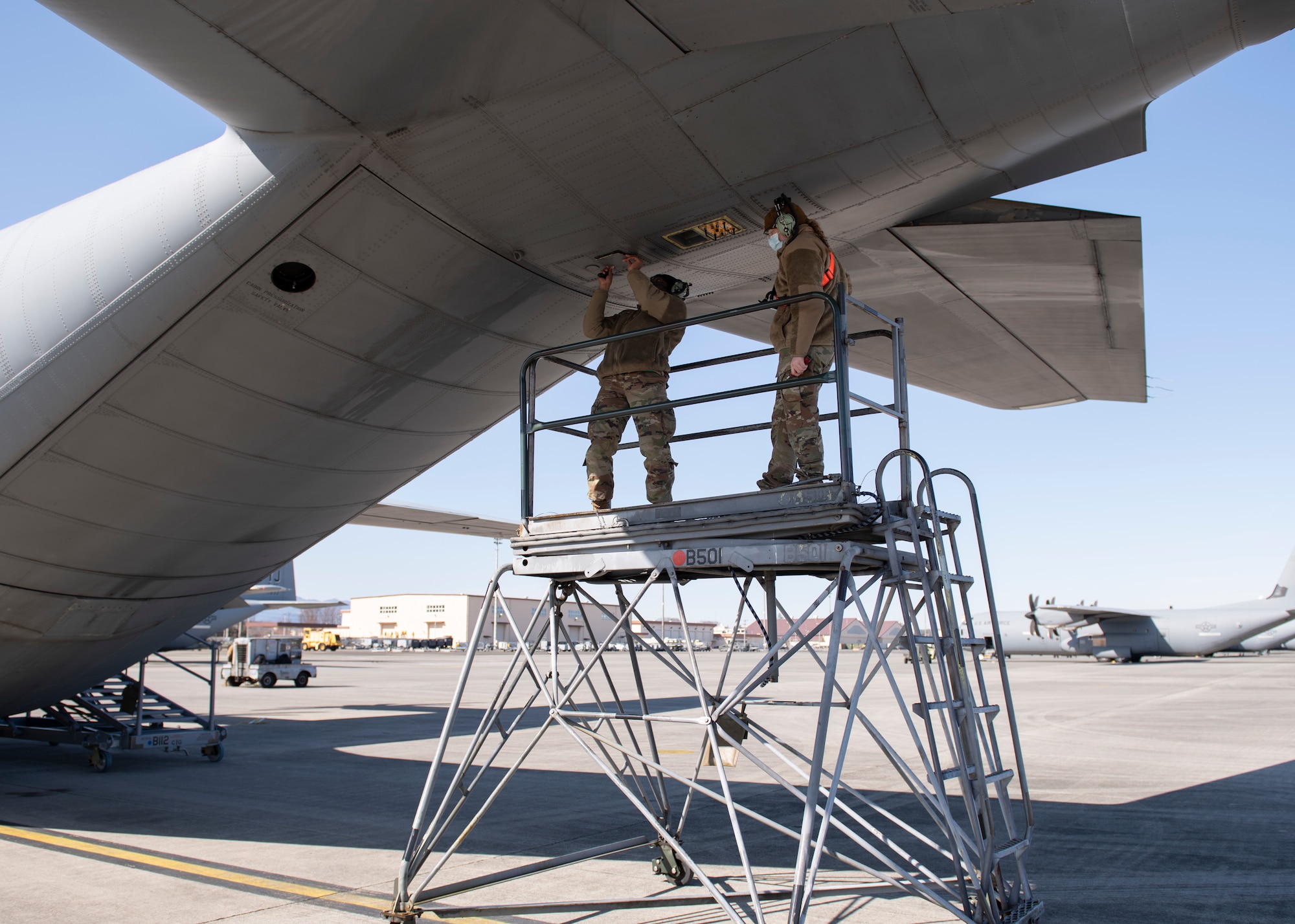 Two airmen balance on a lift as they install chaff and flare cartridges into the underside of a military cargo plane's tail.