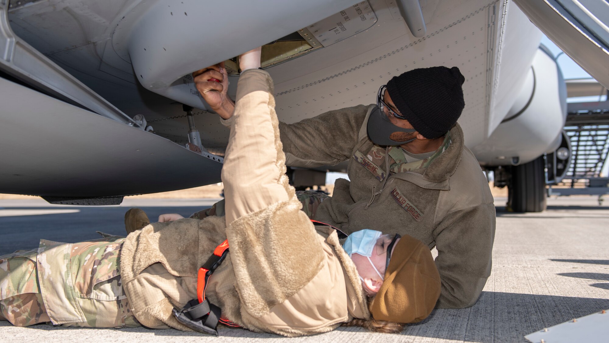 Two airmen lay under the nose of a military cargo plane and test firing pins with a voltmeter.