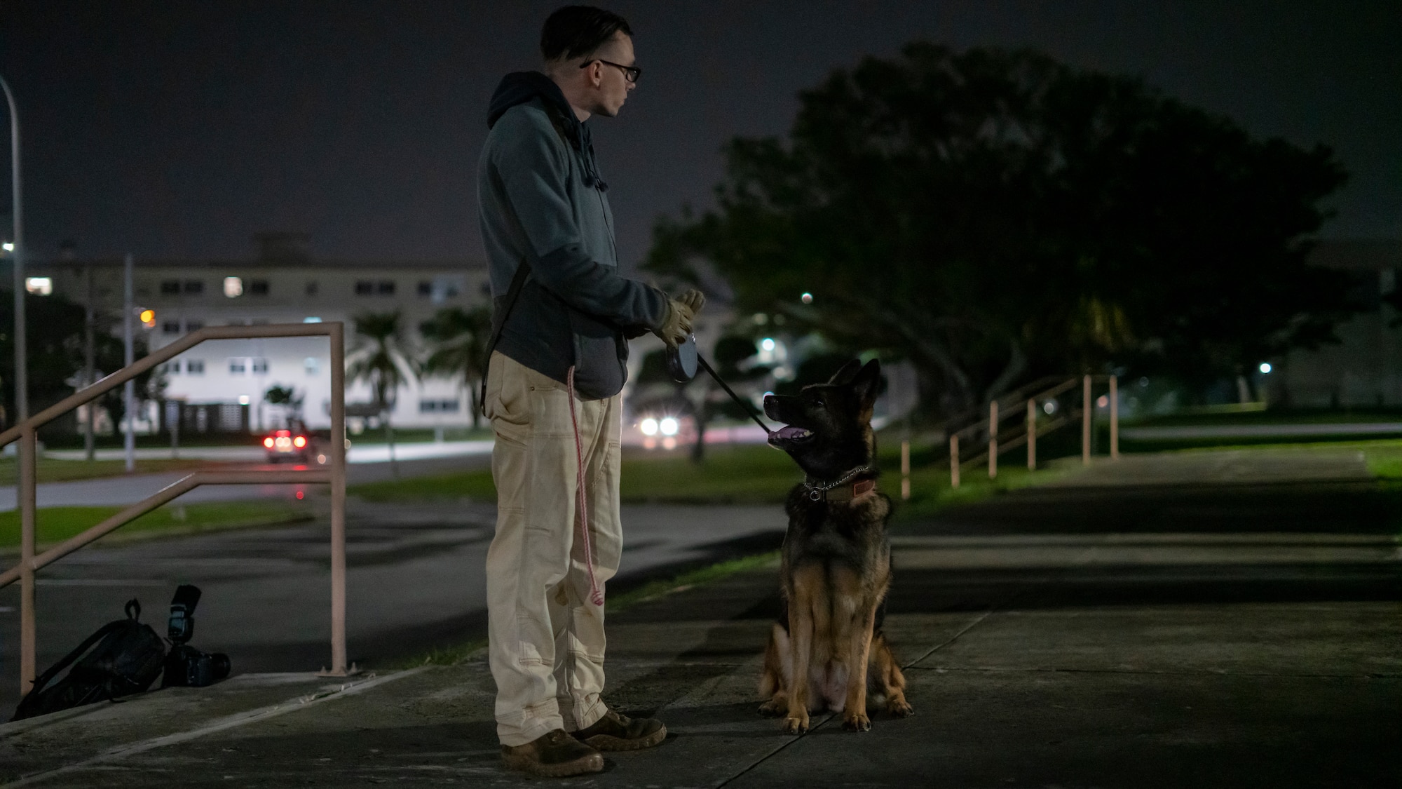 Military working dog trainer stands alongside his dog as they prepare to inspect a building for simulated explosives