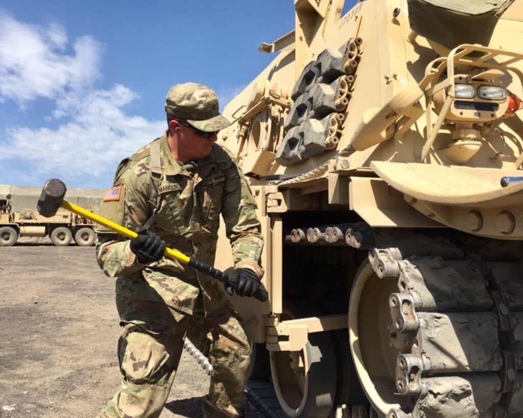 A soldier in a OCP Army uniform with a sledge hammer about to perform maintenance on a tan tank-like wheeled vehicle.