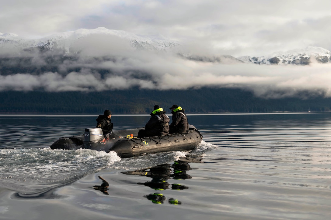 Soldiers in a small rubber boat transit a body of water with snowy mountains in the background.