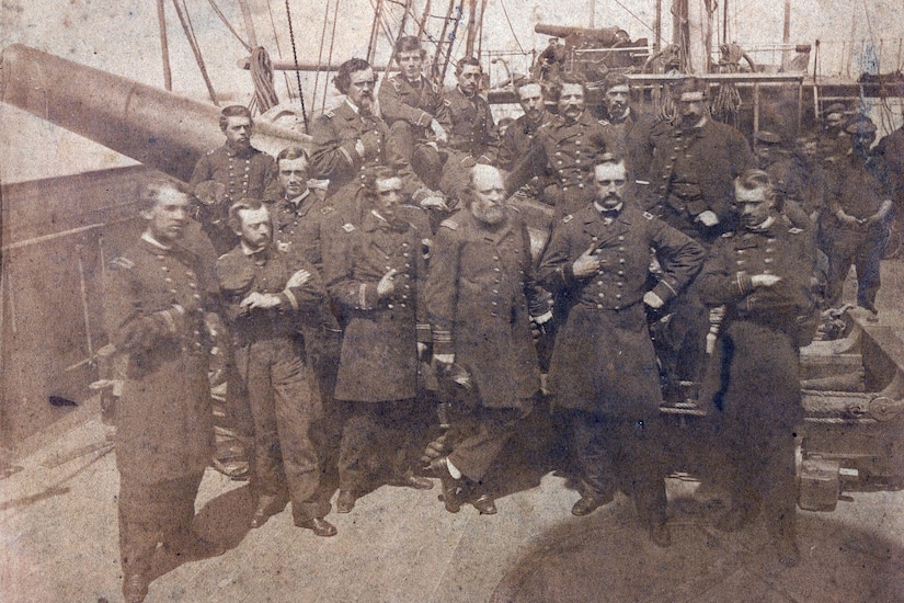 More than a dozen men in uniform pose on a ship’s deck for a photo.