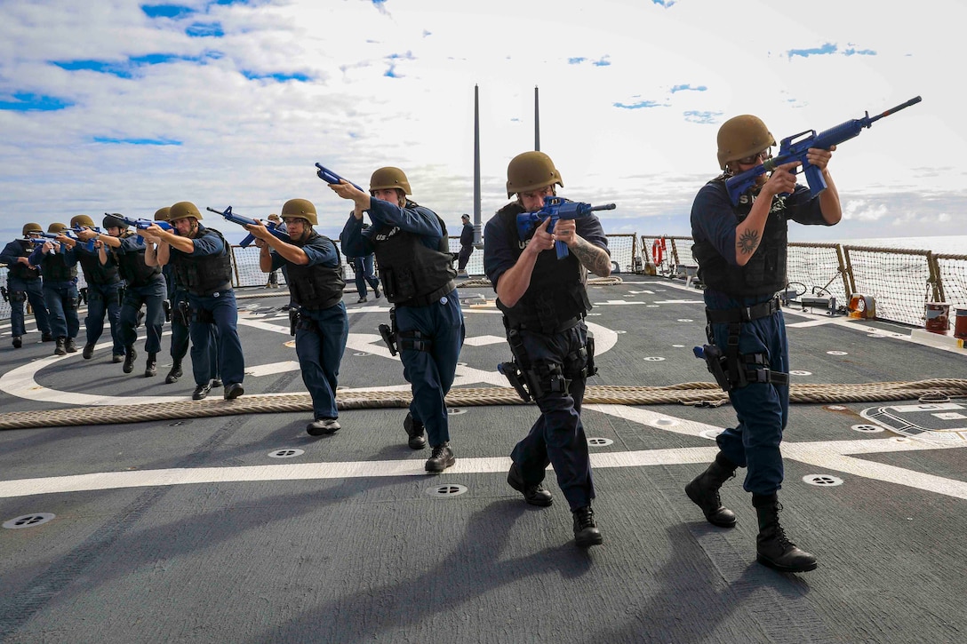 Sailors walk in a line while holding weapons.