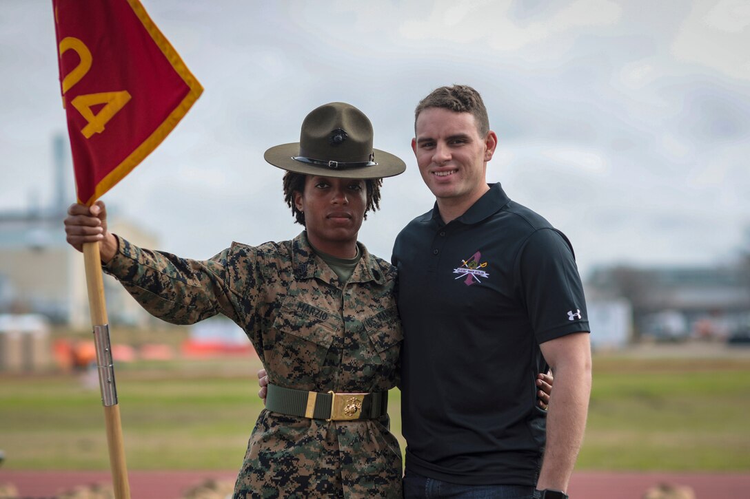 Two Marines stand for a photo outside, one in civilian clothes and the other holding a guidon.