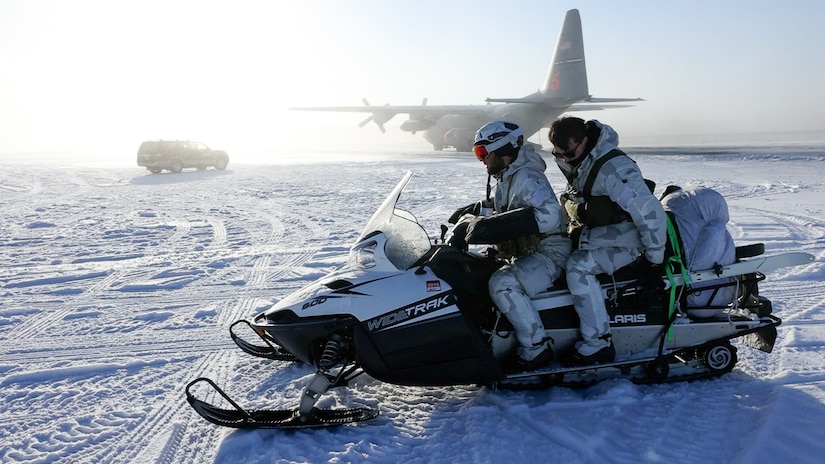 Two individuals sit on a snowmobile. In the background is an airplane.
