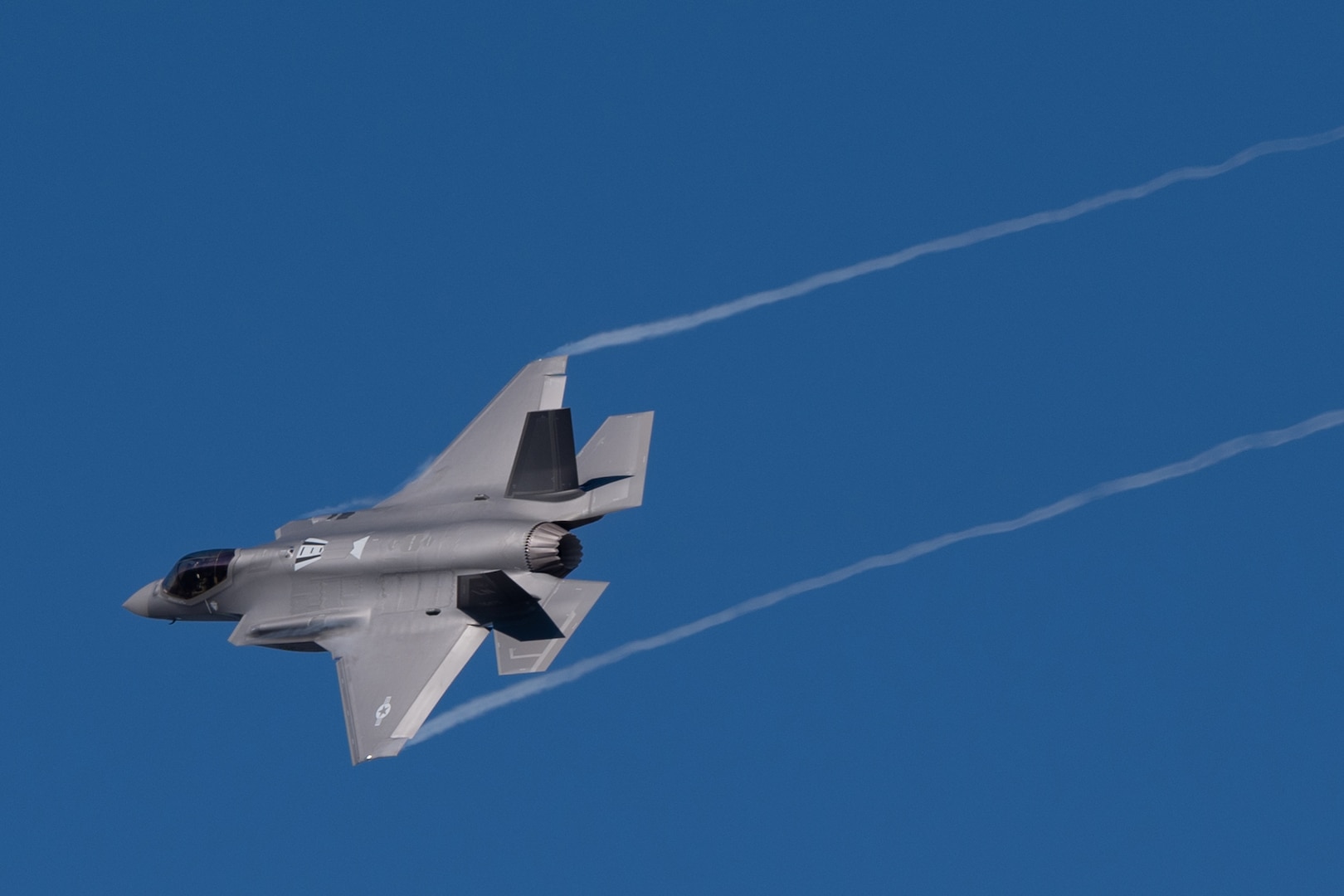 A military aircraft flies across a blue sky creating condensation trails from its wingtips.