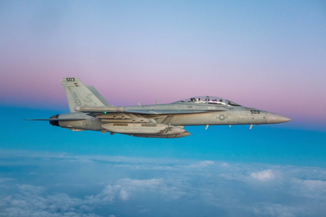 A fighter jet flies across a colorful sky.