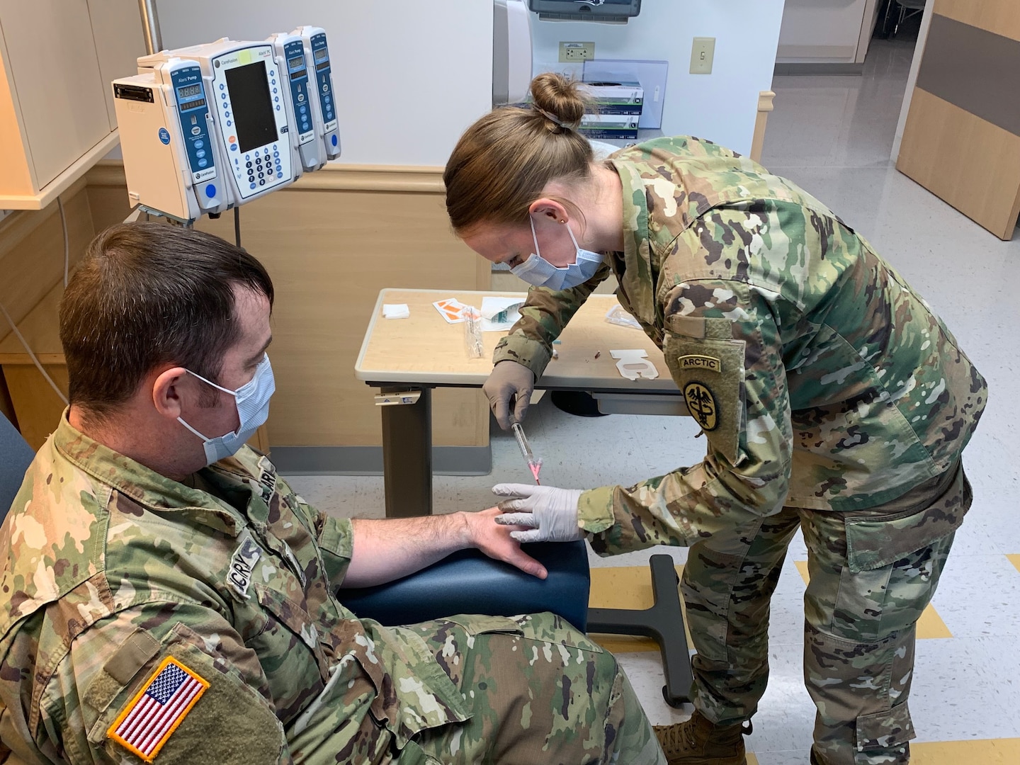 Female and male soldier in uniform. Female soldier starting an IV on the male soldier sitting down.