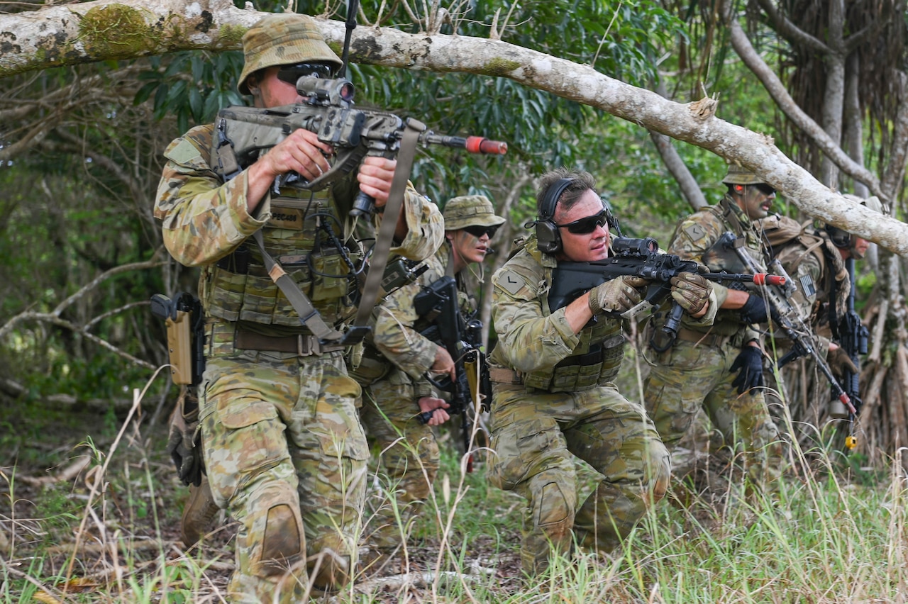 Five service members, wearing camouflage uniforms and carrying weapons, maneuver through brush and trees.