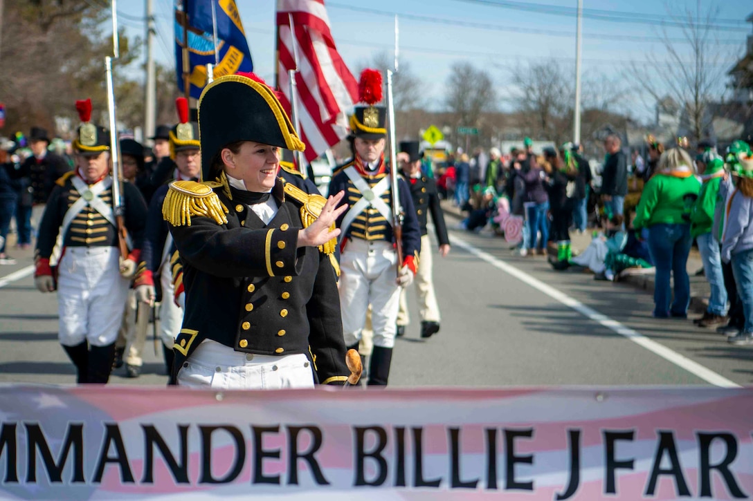 A sailor waves to a crowd as other sailors march behind.