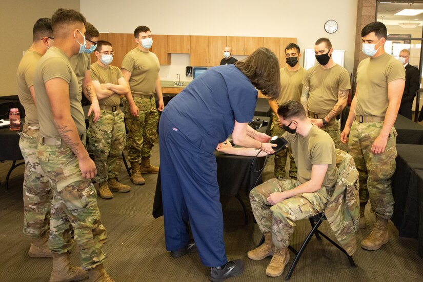 A group of soldiers gather around a woman who is demonstrating how to check a person's blood pressure.