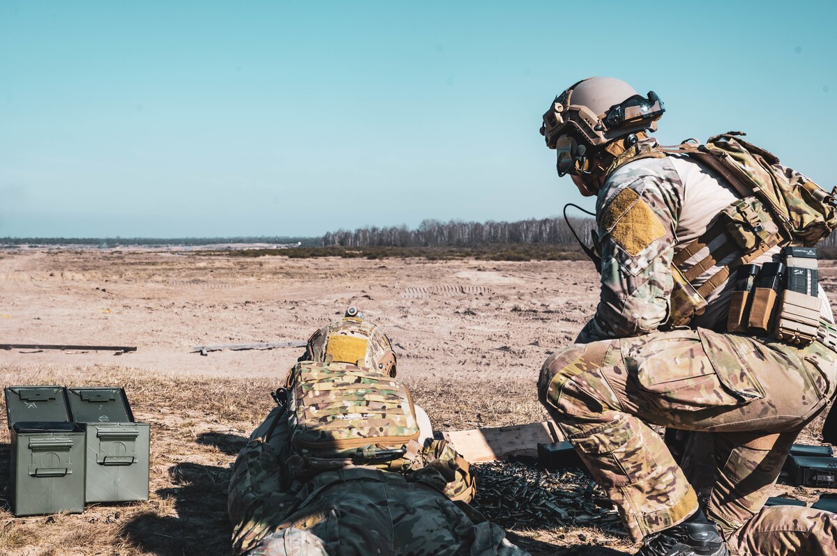An airman shoots a weapon into a barren field as another airman looks on.