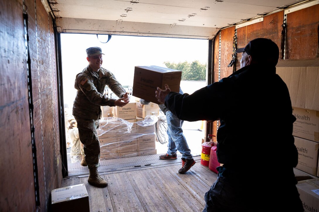 A soldier catches a box of COVID-19 relief supplies being unloaded from a truck. Another man helps to unload the boxes.