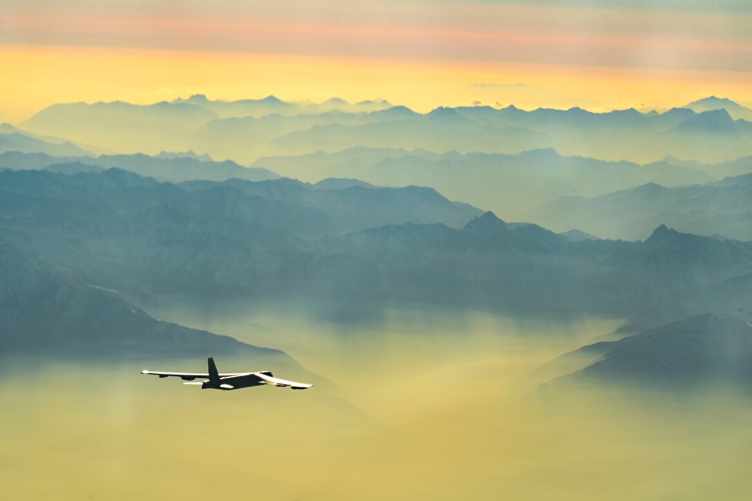 An aircraft flies above mountains near a pink and yellow sky.