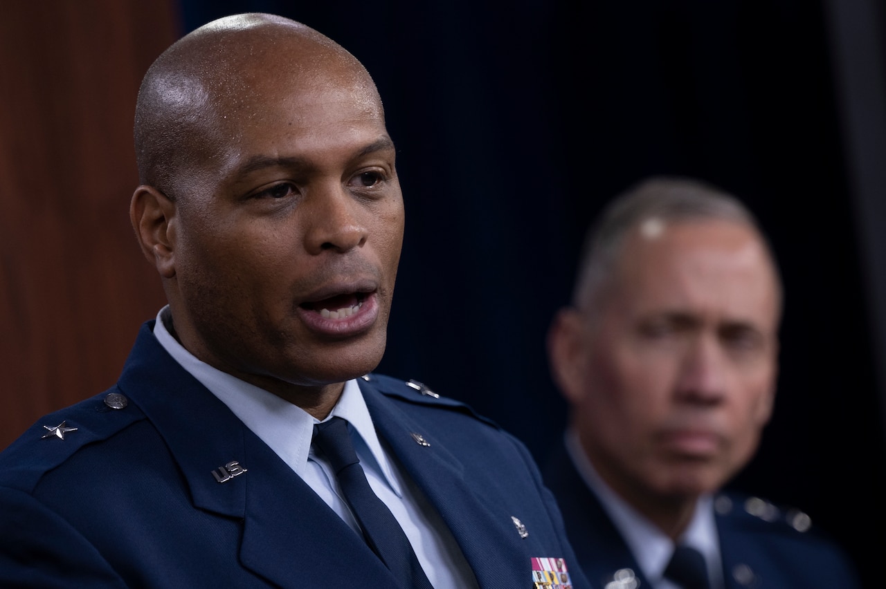 A man in military uniform speaks. Another man in the background looks on.