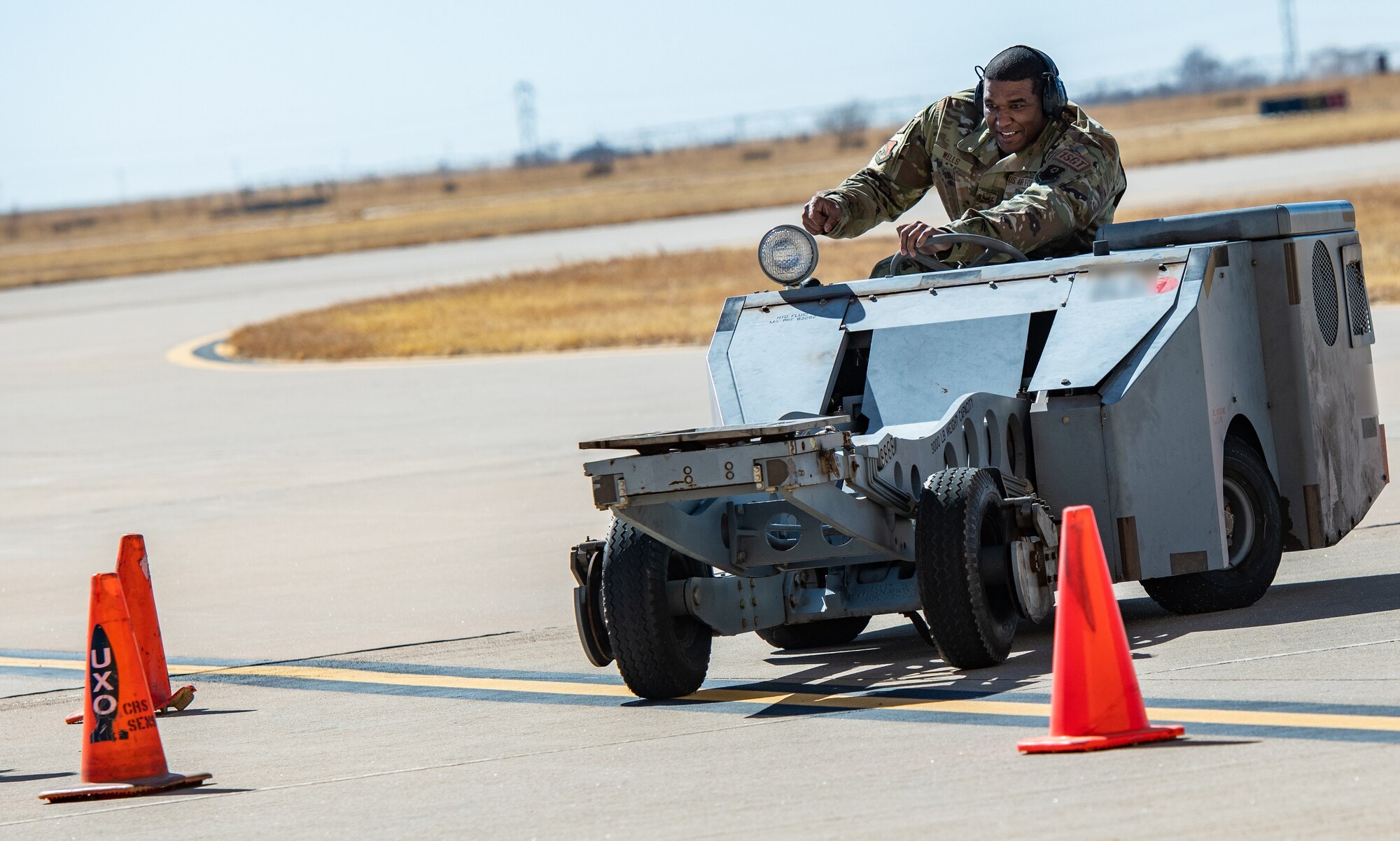 An Air Force sergeant smiles and points at a traffic cone as a drives a bomb lift truck.