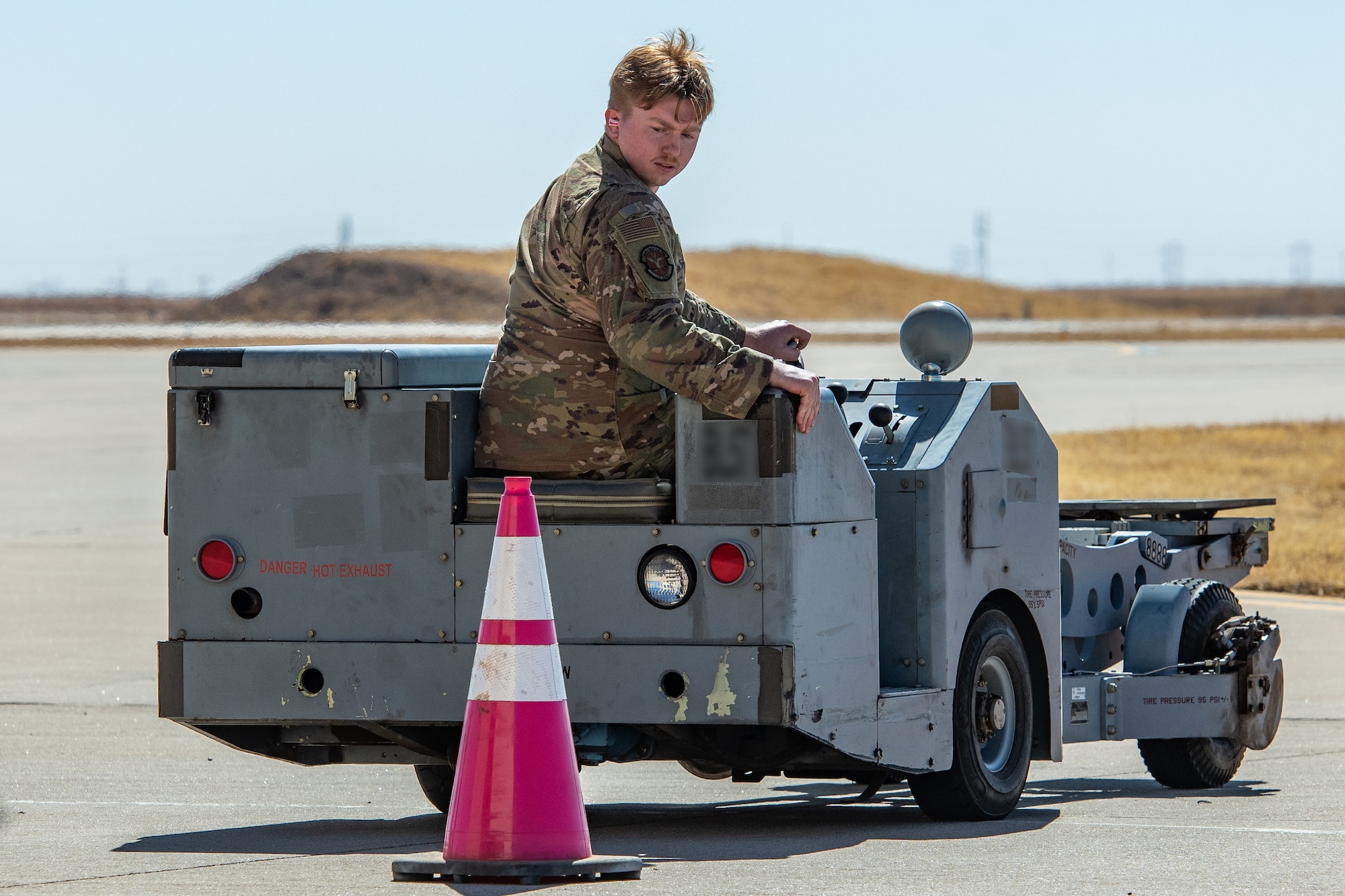 An Airman driving a bomb lift cart looks behind him at cone in the way of the cart.