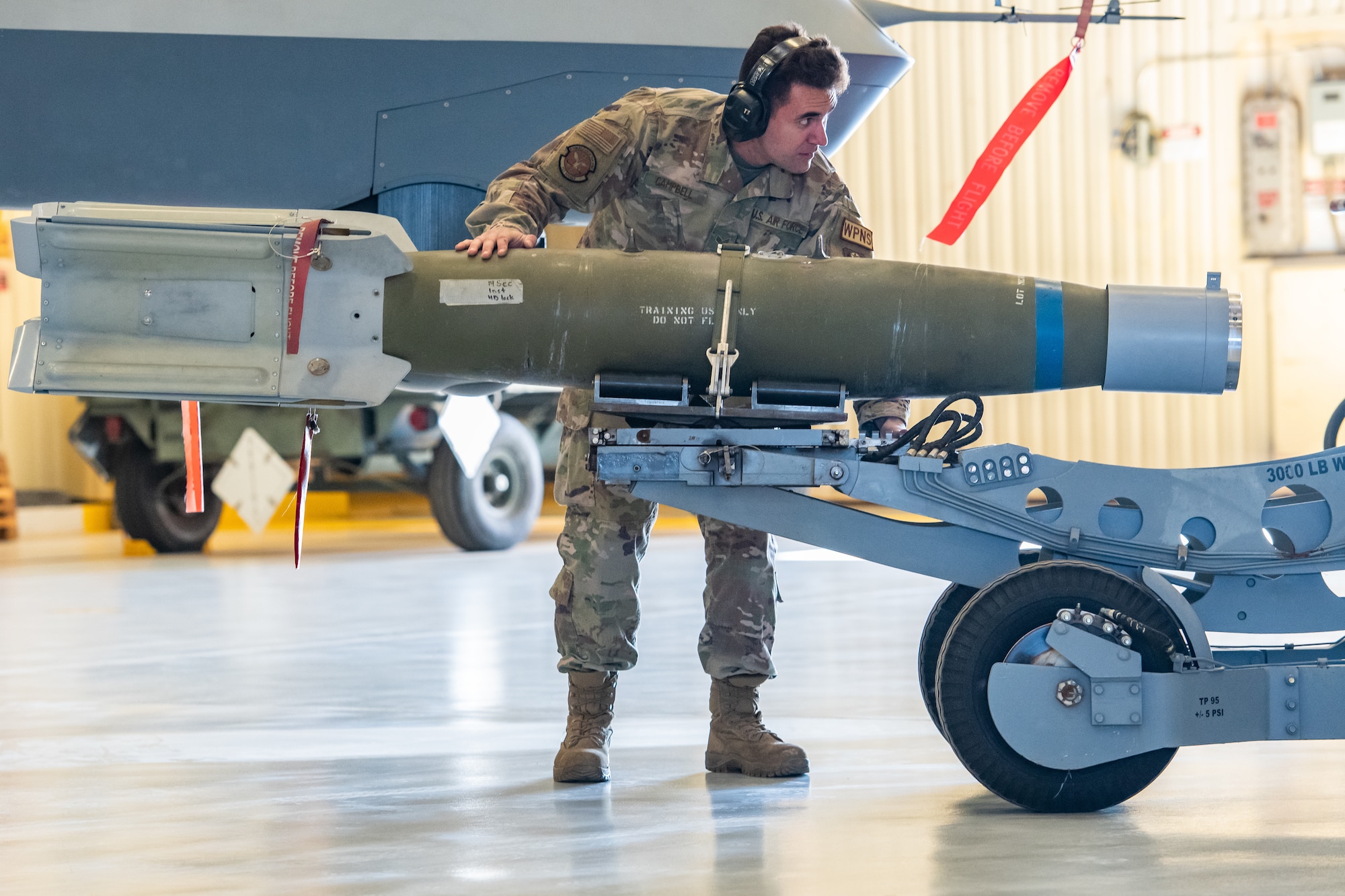 An Airman stand on the side of a bomb on a bomb lift cart and steadies it with his hand.