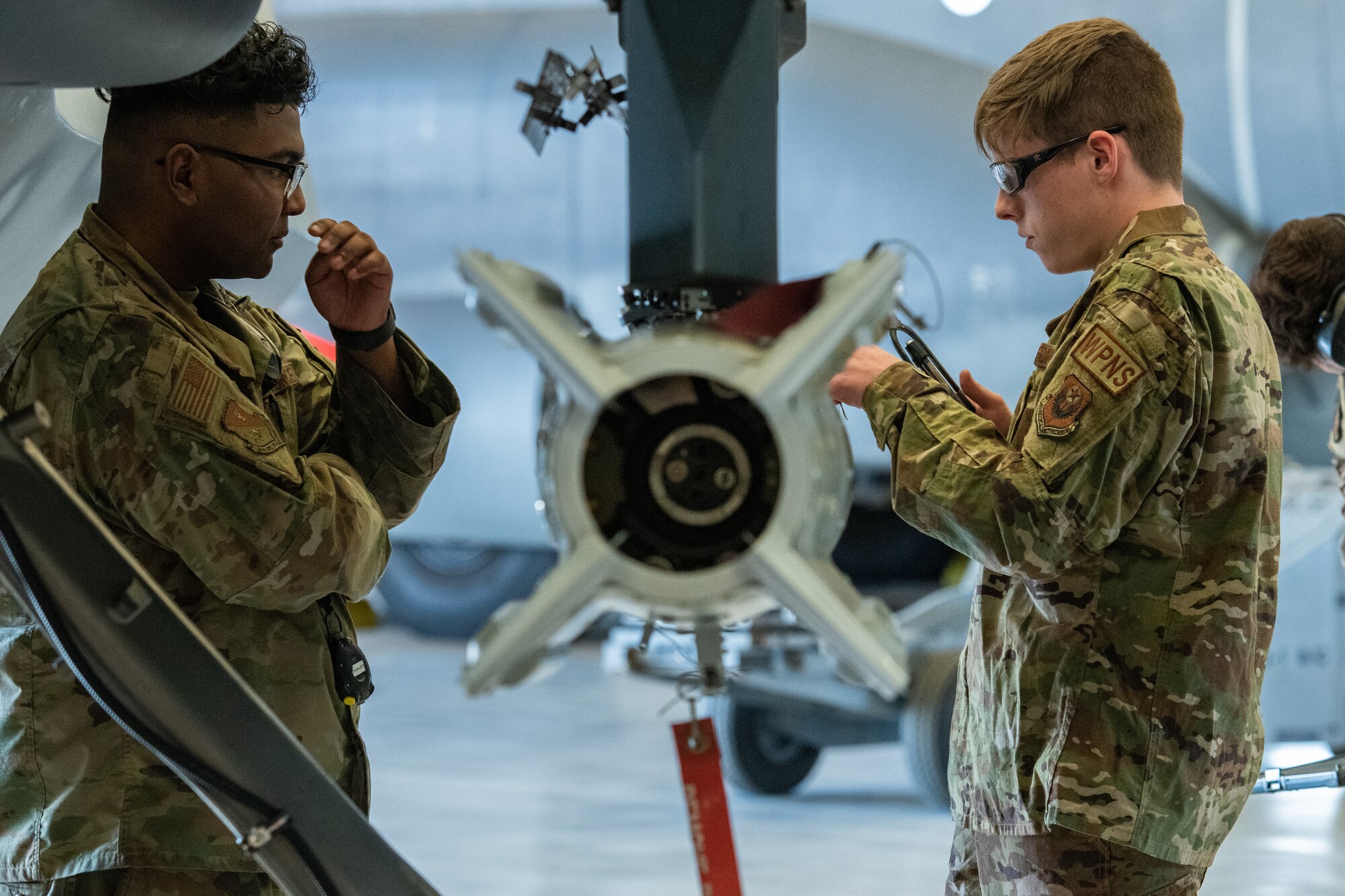 An Airman works on a bomb attached to the wing of a remotely piloted aircraft while another Airman looks on.