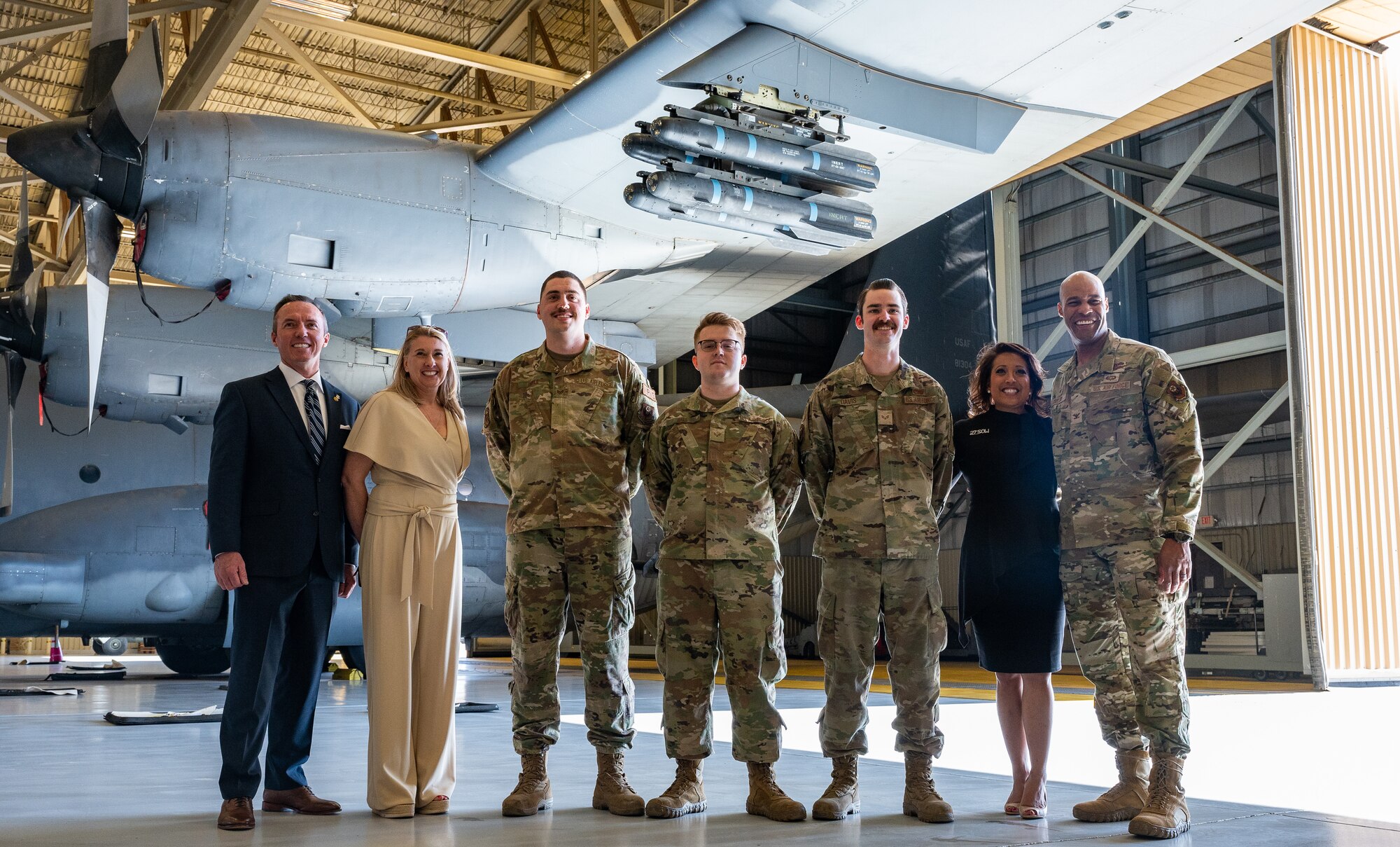 A group of Airmen, a man in a business suit and two women in business attire pose for a group photo in front of missiles hung on a turboprop cargo aircraft wing.
