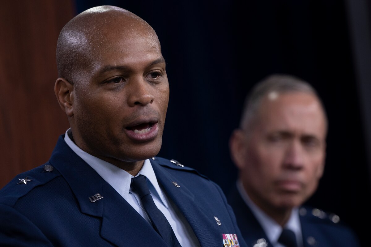 A man in military uniform speaks. Another man in the background looks on.