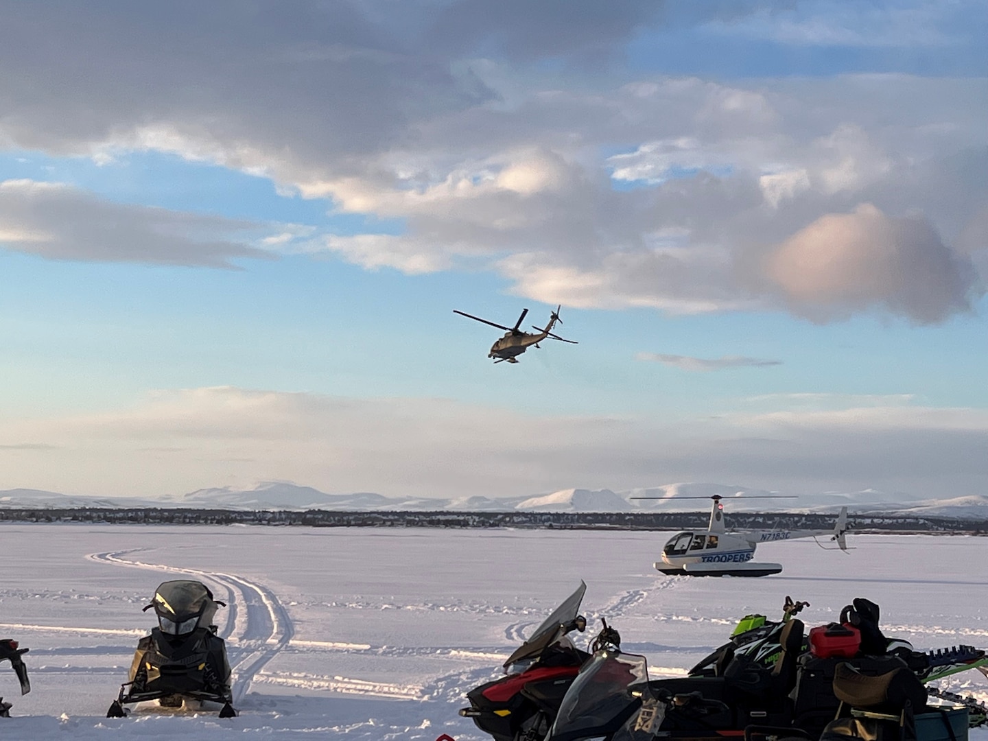 A 210th Rescue Squadron HH-60G Pave Hawk helicopter conducts rescue operations March 5, 2022, at the Lake Iliamna crash site of a Cessna 206. On the ground are an Alaska State Trooper R-44 helicopter and good Samaritans’ snowmachines in the foreground.
