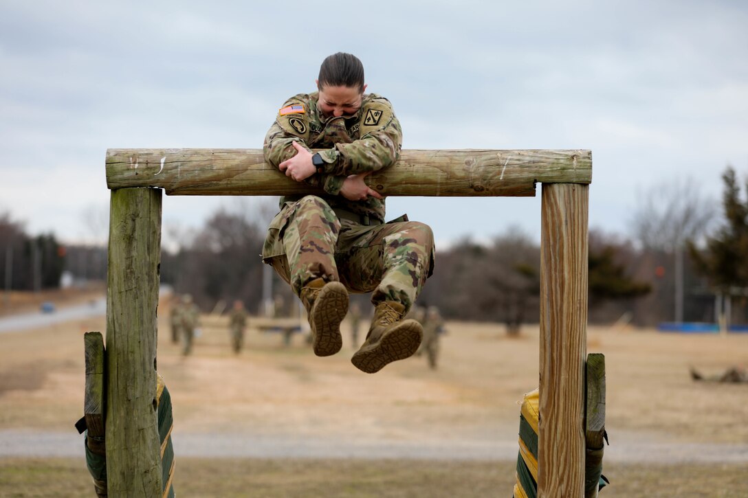 A soldier grimaces as she climbs over a high wooden bar.