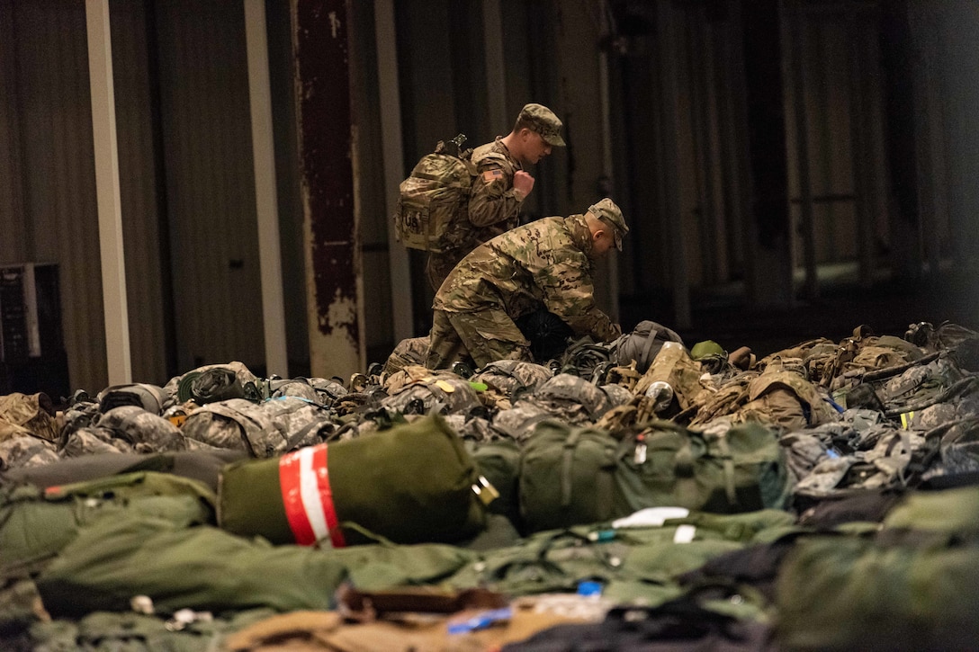 Two soldiers work with their gear in a large room covered with equipment.