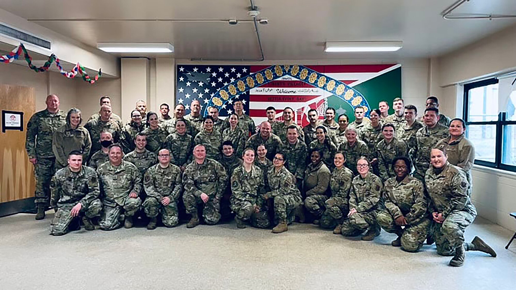 Group photo of about 40 United States Air Force members posing before an "Operation Allies Welcome" banner. The banner depicts a gateway, the U.S. flag and the Afghanistan flag.