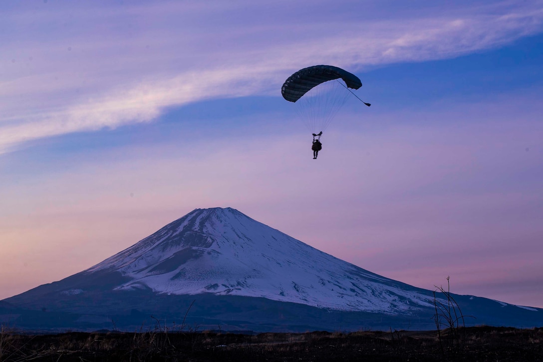 A Marine descends in the sky wearing a parachute; a mountain can be seen in the background.