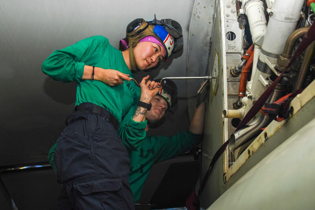 A sailor uses a tool to work on an aircraft as another sailor watches.