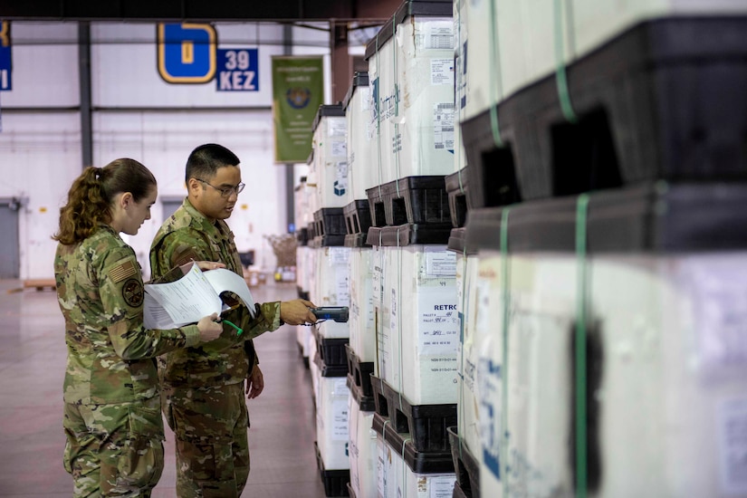 A male service member holds a scanner up to one of many crates stacked in a large warehouse, which a female service member looks at papers on a clipboard.