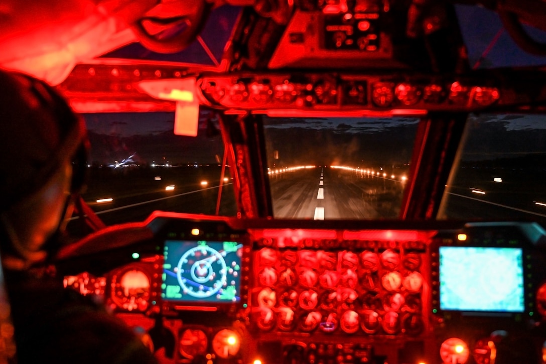An airman in a cockpit lands a large plane on a lit runway at night.