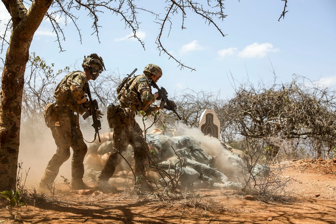 Two soldiers hold weapons and move forward on dirt near sandbags and a target.