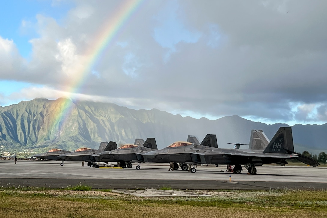 Aircraft sit on a flightline with a rainbow and mountains in the background.