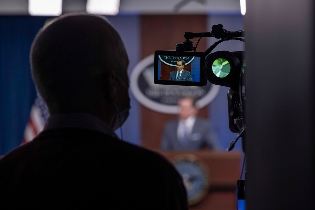 A person holds up a video camera, which displays on its screen the image of a person briefing reporters.