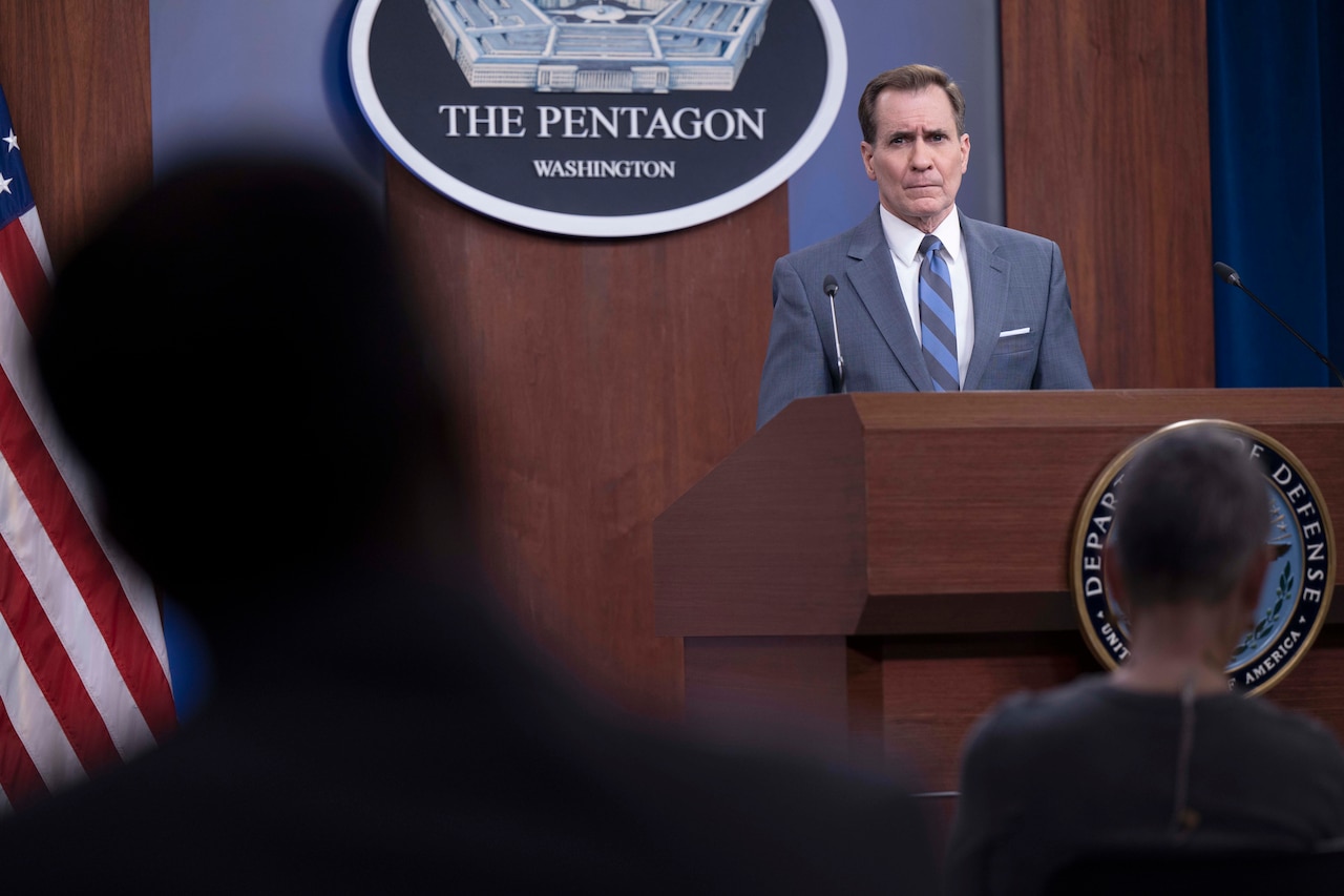 A civilian speaks at a lectern to a seated audience.