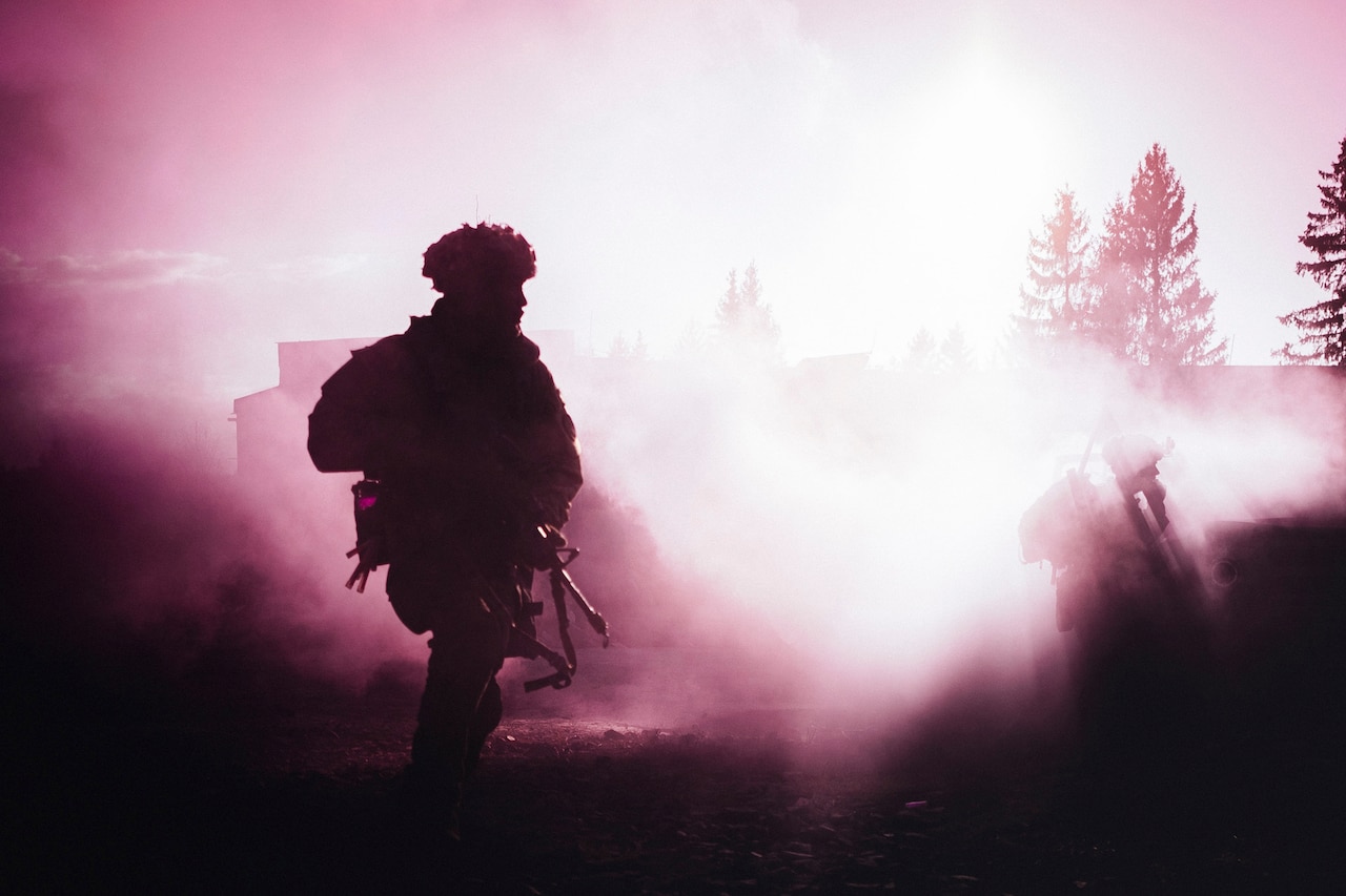 A soldier holding a weapon walks through smoke in a forest.