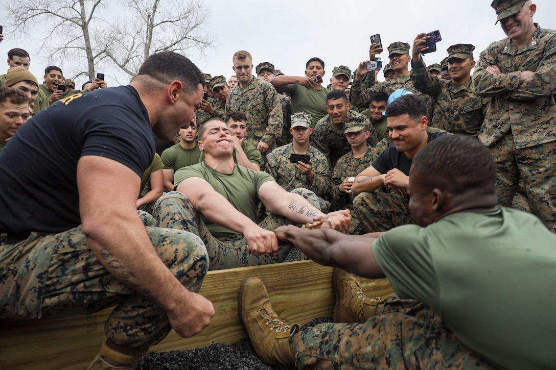 Marines facing each other pull a stick simultaneously while placing their feet on a board while fellow Marines watch.