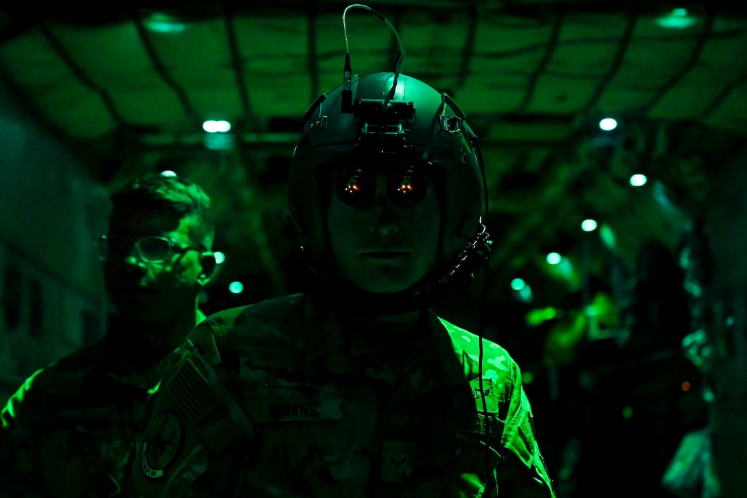 An airman illuminated by green light wears a helmet and googles as another airman stands beside.