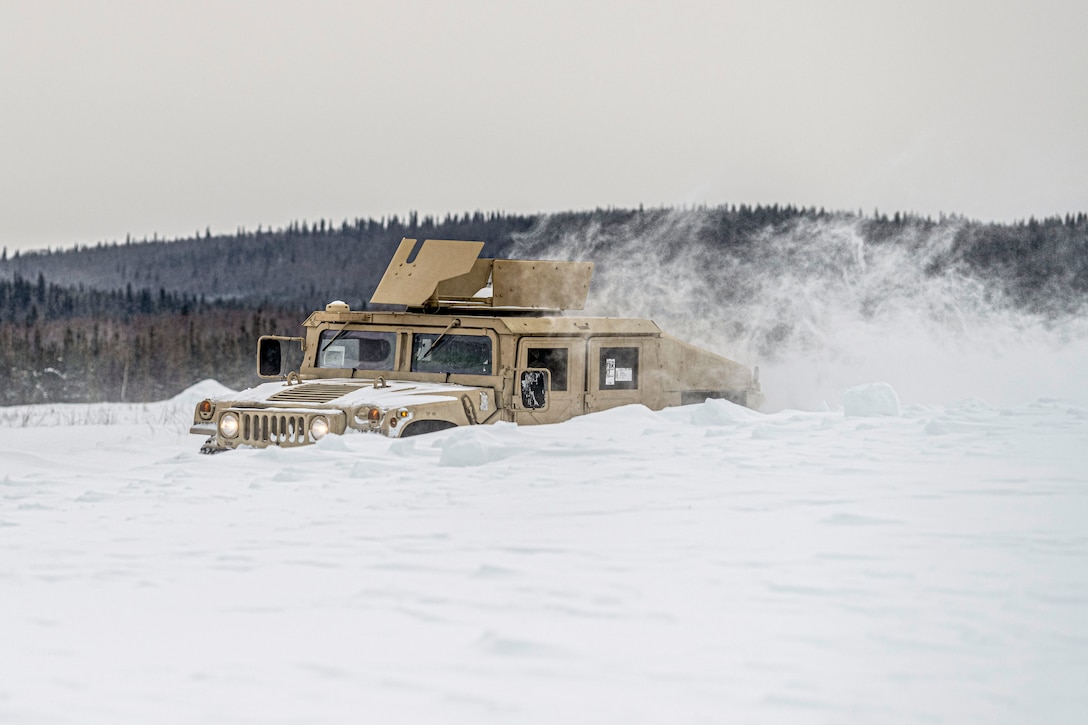 An Army Humvee drives through snow almost up to its headlights, leaving a powdery cloud in its wake.