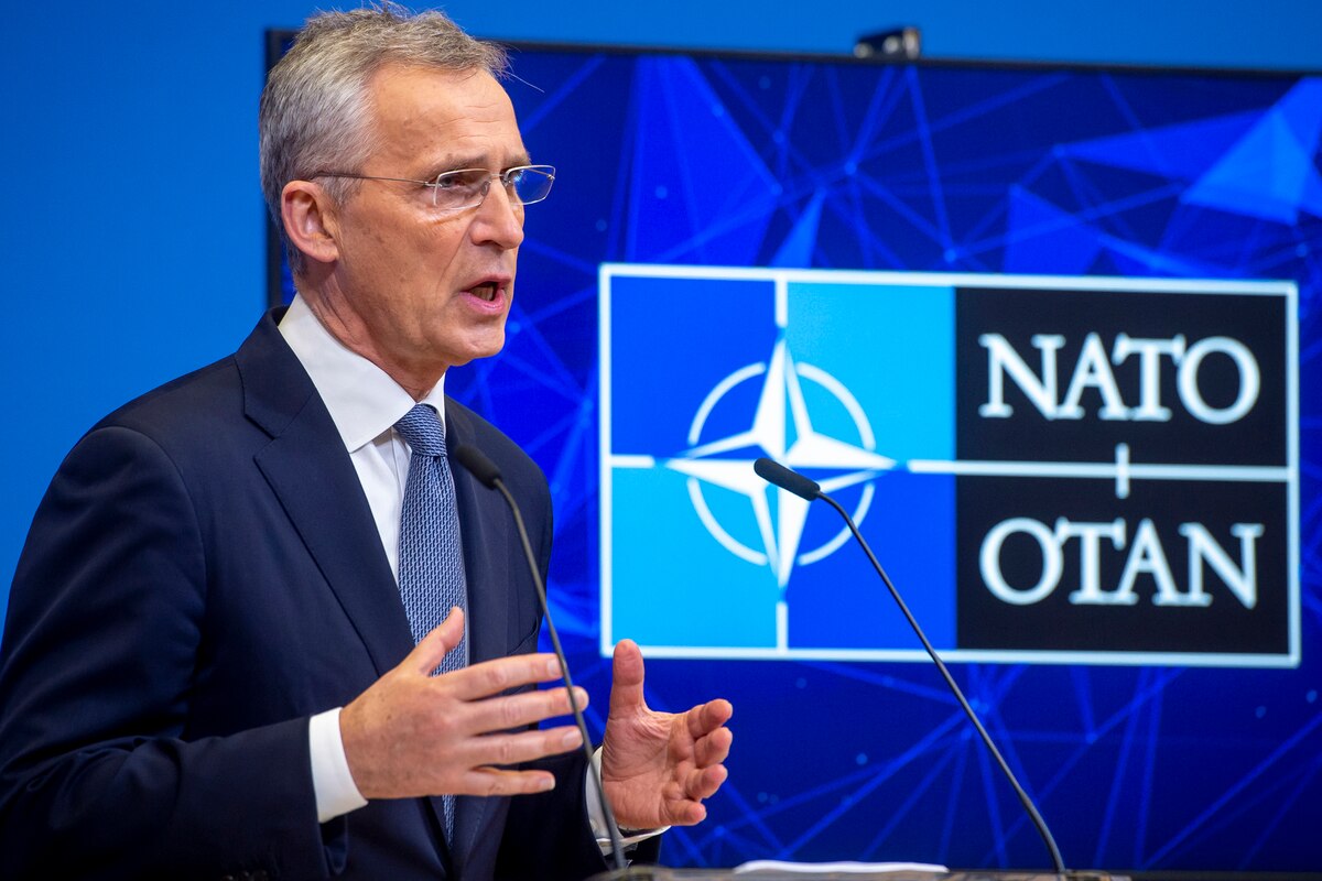 A man in a suit stands behind a lectern. Behind him are signs that say "NATO."
