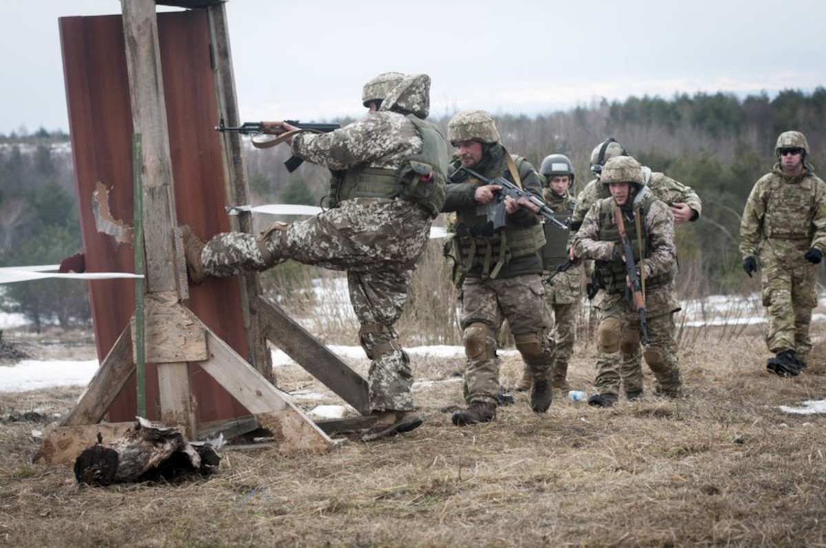 Uniformed military personnel carry rifles outdoors. One kicks open a free-standing door.