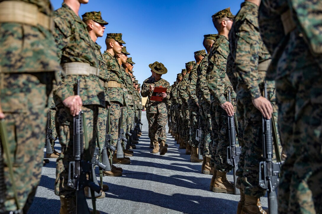 Marines holding clipboards walk between rows of Marines at attention.