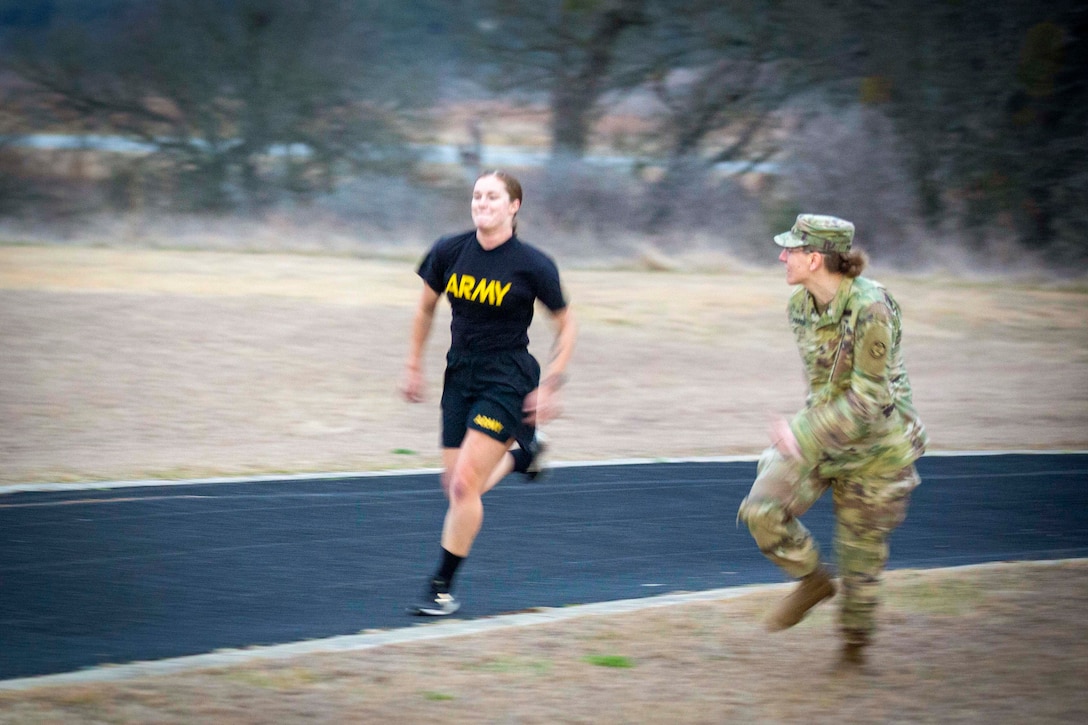 A soldier cheers on a fellow soldier as she runs on a track.