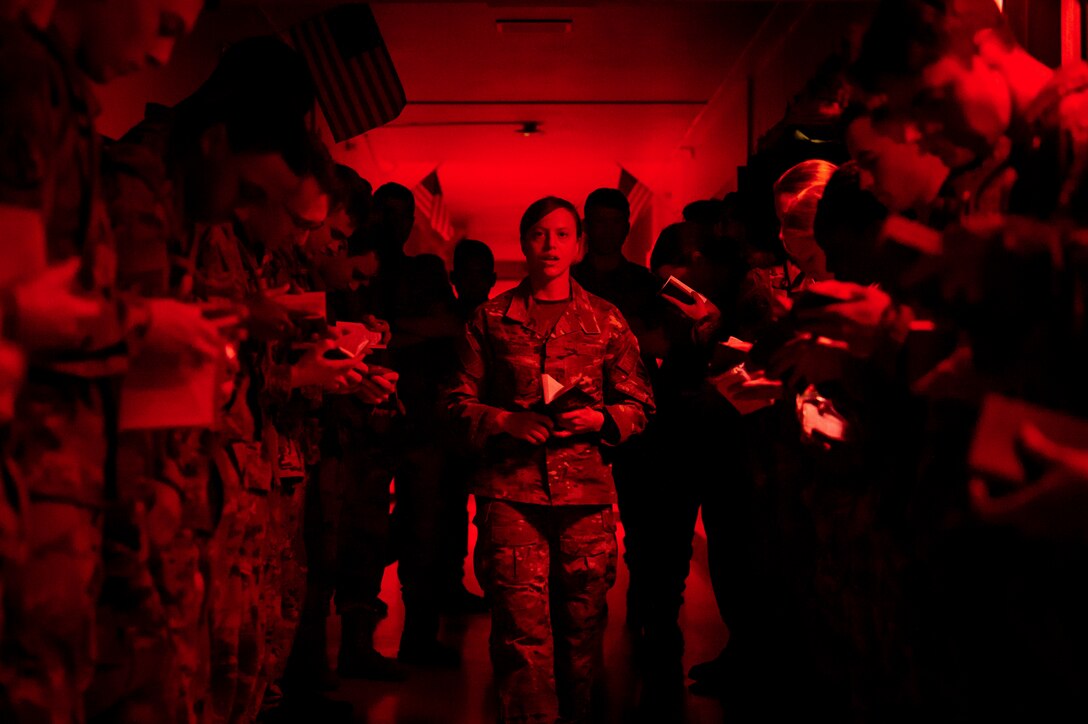 Air Force cadets read from small books while standing in two parallel lines as airmen walk down the center illuminated by red light.