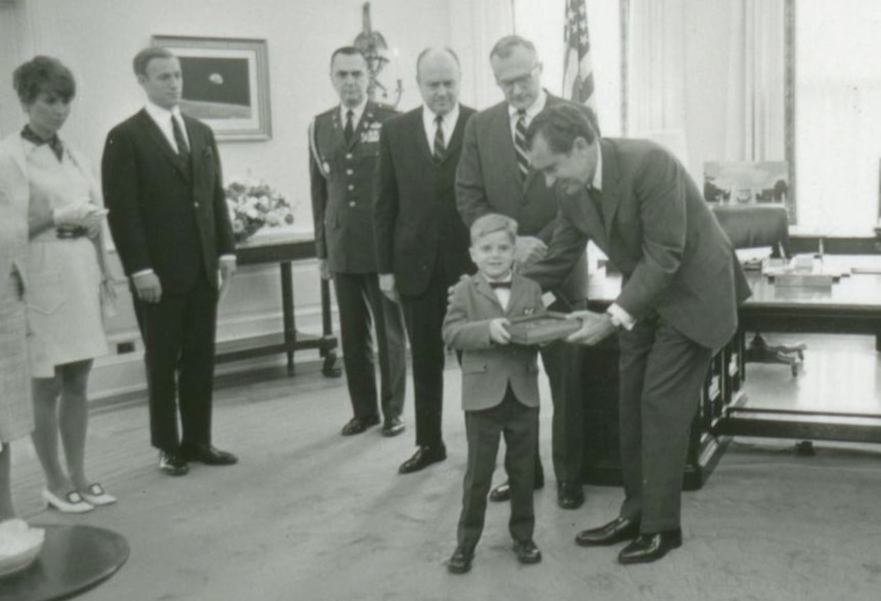 A man puts his arm around a boy’s shoulder as they each hold a small wooden box. Several people stand around them