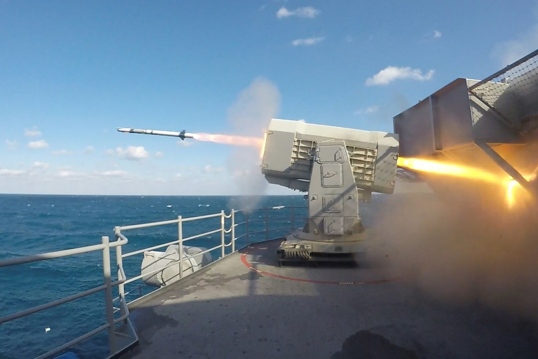 A missile shoots flames as it fires from a ship's deck over blue water.